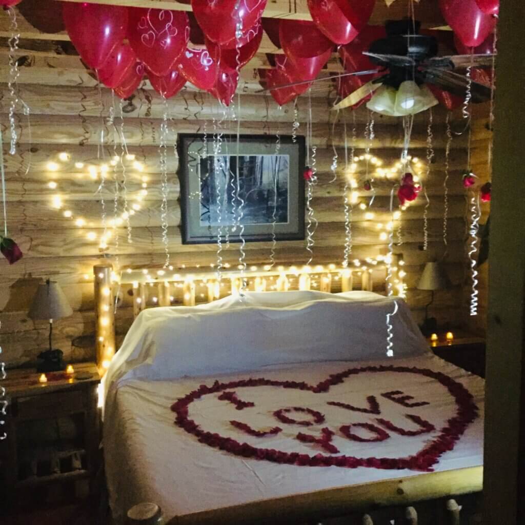 Romantic cabin bedroom decorations with balloons on the ceiling, hearts on the wall, lights on the bed, and I Love You message in rose petals on the bed